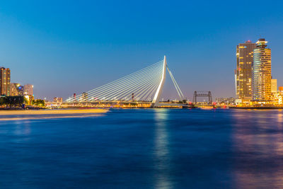 Illuminated erasmus cable-stayed bridge over the river maas by buildings against clear blue sky