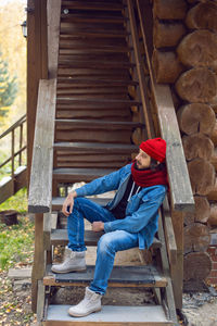Man with a hipster beard in red knitted hat and, a denim jacket is sitting on stairs wooden house