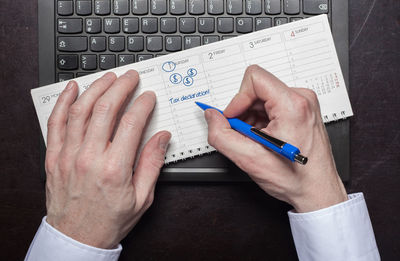 Cropped hands of businessman writing in book on laptop at desk
