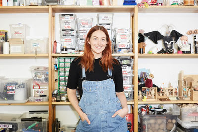 Portrait of confident redhead female technician standing against shelf at workshop