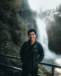 Portrait of young man standing against waterfall in forest