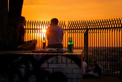 Rear view of people sitting on railing against orange sky