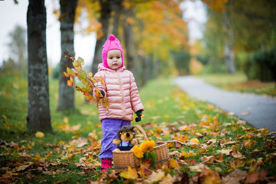Cute girl holding dry twig standing by wicker basket with stuffed toy at roadside
