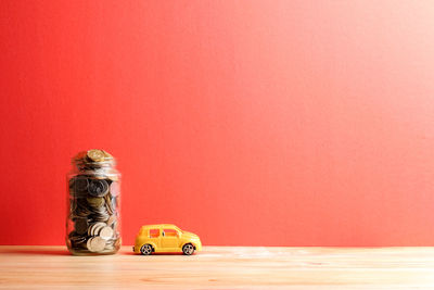 Coins in jar with toy car on wooden table against wall