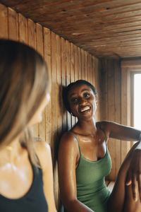 Happy young woman talking with female friend while sitting in wooden sauna