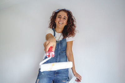 Portrait of smiling woman holding paint roller while standing against wall