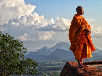 Monk looking away while standing on retaining wall against sky