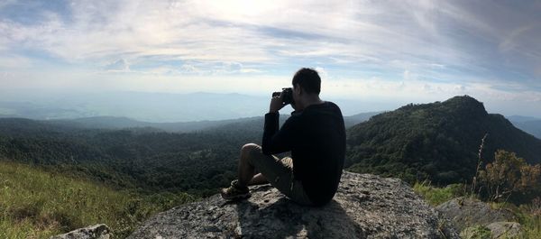 Man photographing while sitting on cliff against cloudy sky