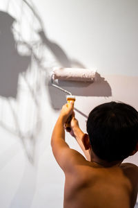 Rear view of shirtless boy holding paint roller on wall