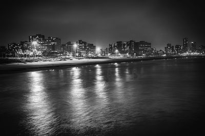 Illuminated buildings by sea against sky at night