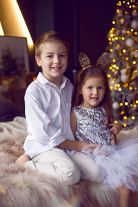 Children sister and brother sitting on the bed on new year's eve next to the tree at christmas