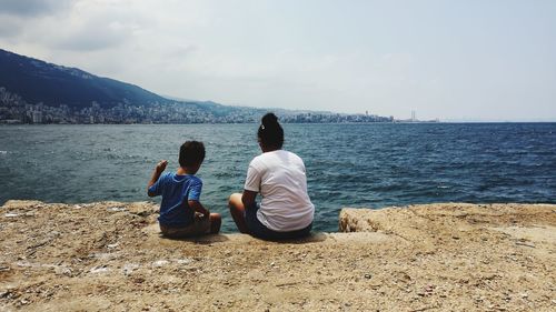 Rear view of siblings sitting at beach against sky