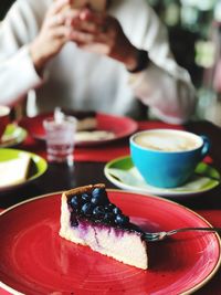 Close-up of cake slice in plate with person using mobile phone in background on table