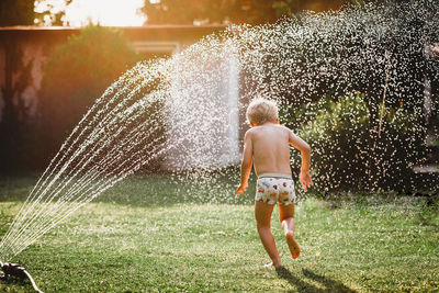 Young white boy running under the water from the sprinkler in garden