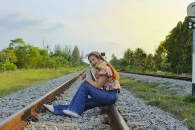 Full length portrait of woman sitting on railroad track against sky
