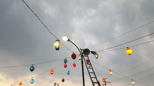 Low angle view of illuminated lanterns hanging on cables against cloudy sky