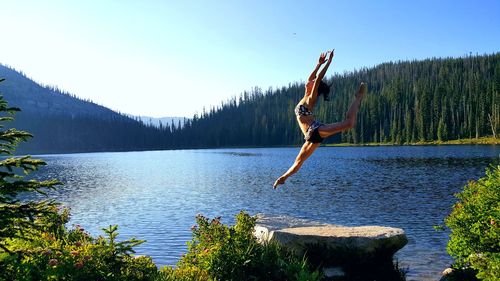 Full length of woman jumping by river with arms raised against sky