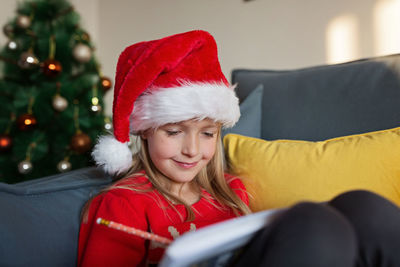 Cute blonde caucasian girl 7 years old sitting on couch near christmas tree writing letter to santa