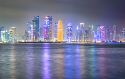 Illuminated buildings by sea against sky at night