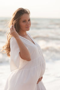 Beautiful calm pregnant woman wear white dress posing over sea at background closeup.