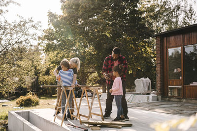 Mother and son using hand saw while father talking to daughter outside house