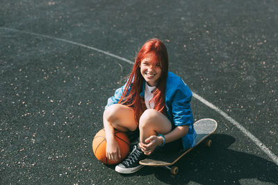 A smiling girl with a basketball is sitting on a skateboard on the sports field