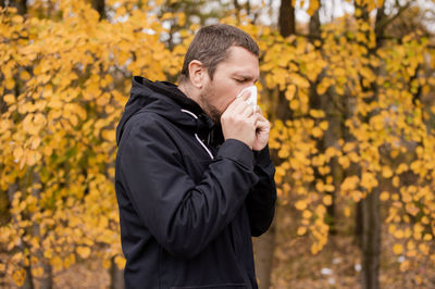 A man in a jacket is standing on the street and holding a napkin near his nose