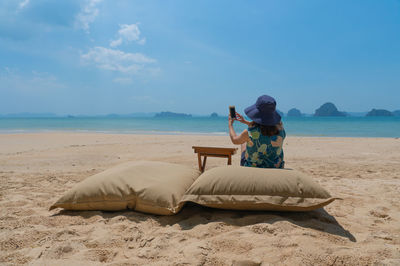 Young woman sitting on bean bag near beach and using smartphone to take photo during summer vacation