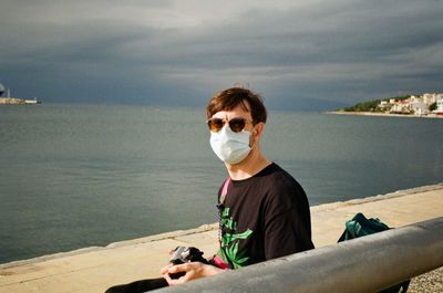 Portrait of man wearing mask and sunglasses sitting against sea