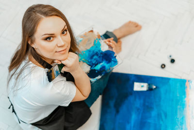 High angle portrait of smiling woman with painting sitting on floor