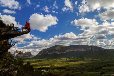 Low angle view of man sitting at the edge of cliff against cloudy sky