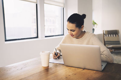 Businesswoman writing while sitting with laptop computer at desk in office