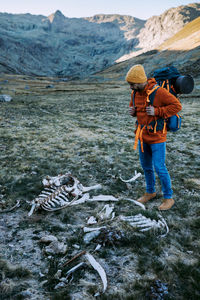 Full body side view of male traveler with backpack observing bones of dead animal while standing on grassy ground against mountain range