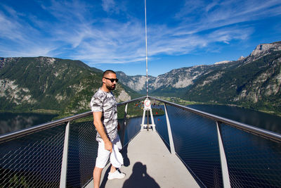 Young woman standing on railing by mountain against sky