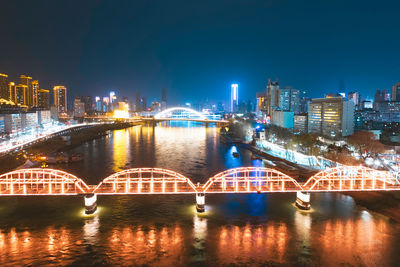 Illuminated bridge over river by buildings against sky at night