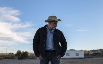 Adult man in cowboy hat standing in front of white building against sky