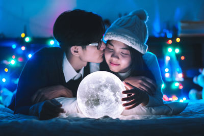 Boyfriend kissing girlfriend by illuminated moon light decoration on bed