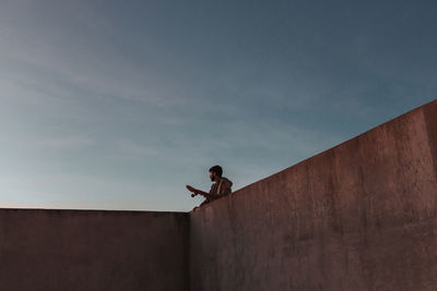 From below of young bearded male skater standing on high concrete ramp in skatepark with skateboard looking away