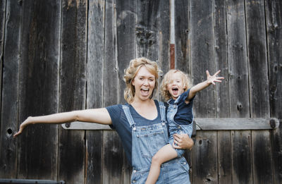 Portrait of cheerful mother and daughter with arms outstretched against old barn