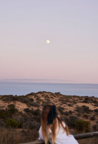 Back view of unrecognizable female tourist supported on wooden fence on embankment and admiring peaceful sunset over sea in summer with the moon in the background