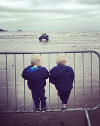 Rear view of boys standing on railing against sea