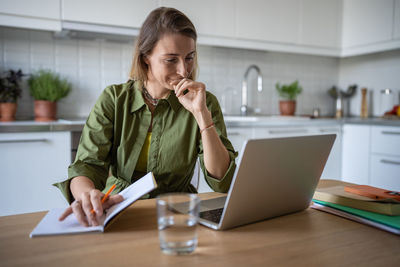 Pensive female working on laptop computer at home, smiling being satisfied with work results