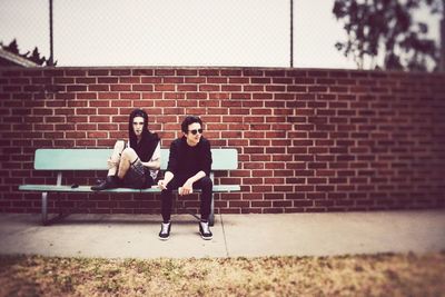 Full length of friends sitting on bench against brick wall