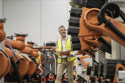 Engineer holding file folder and eyeglasses examining robotic arms in factory