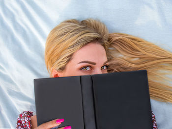 Young blonde woman with green eyes reads a book lying on a blanket, looks out from behind a book