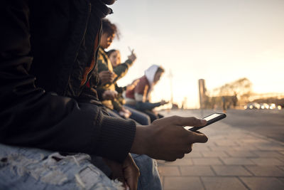 Midsection of teenager using phone while sitting with friends by cobbled street