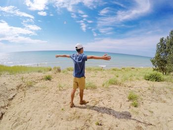 Rear view of young man with arms outstretched standing at beach against sky during sunny day