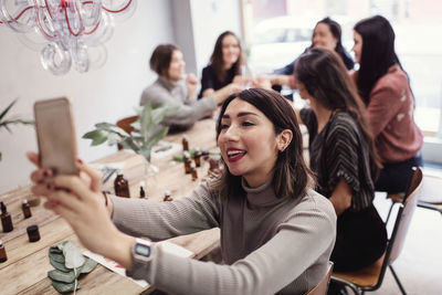 Smiling young woman taking selfie with female colleagues sitting at table in perfume workshop