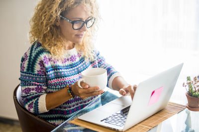 Smiling woman holding coffee cup while working on laptop at home
