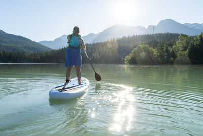 Back view of barefoot woman in dress and life vest riding sup board on calm lake water on sunny summer day in mountains in british columbia, canada